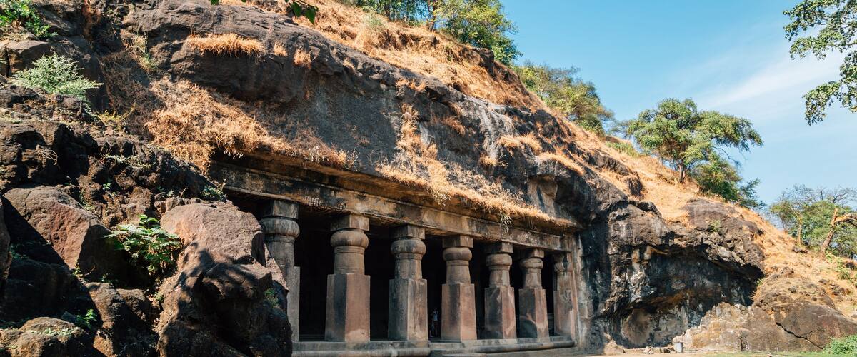 A rock-cut temple with columns, set against a hillside with sparse vegetation and a clear sky.