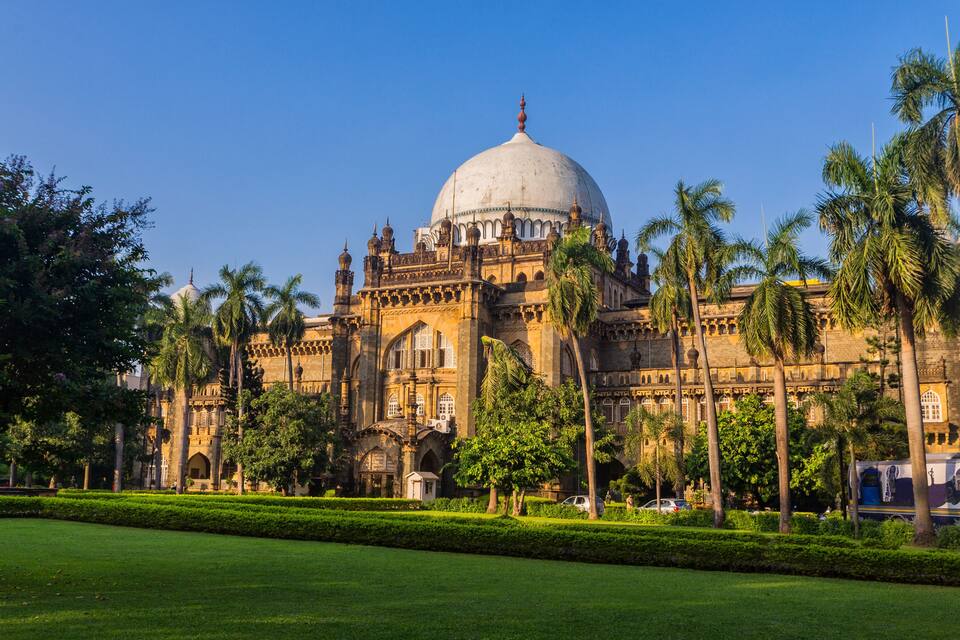 A historic building with a large dome and intricate architecture, surrounded by well-maintained greenery and palm trees.