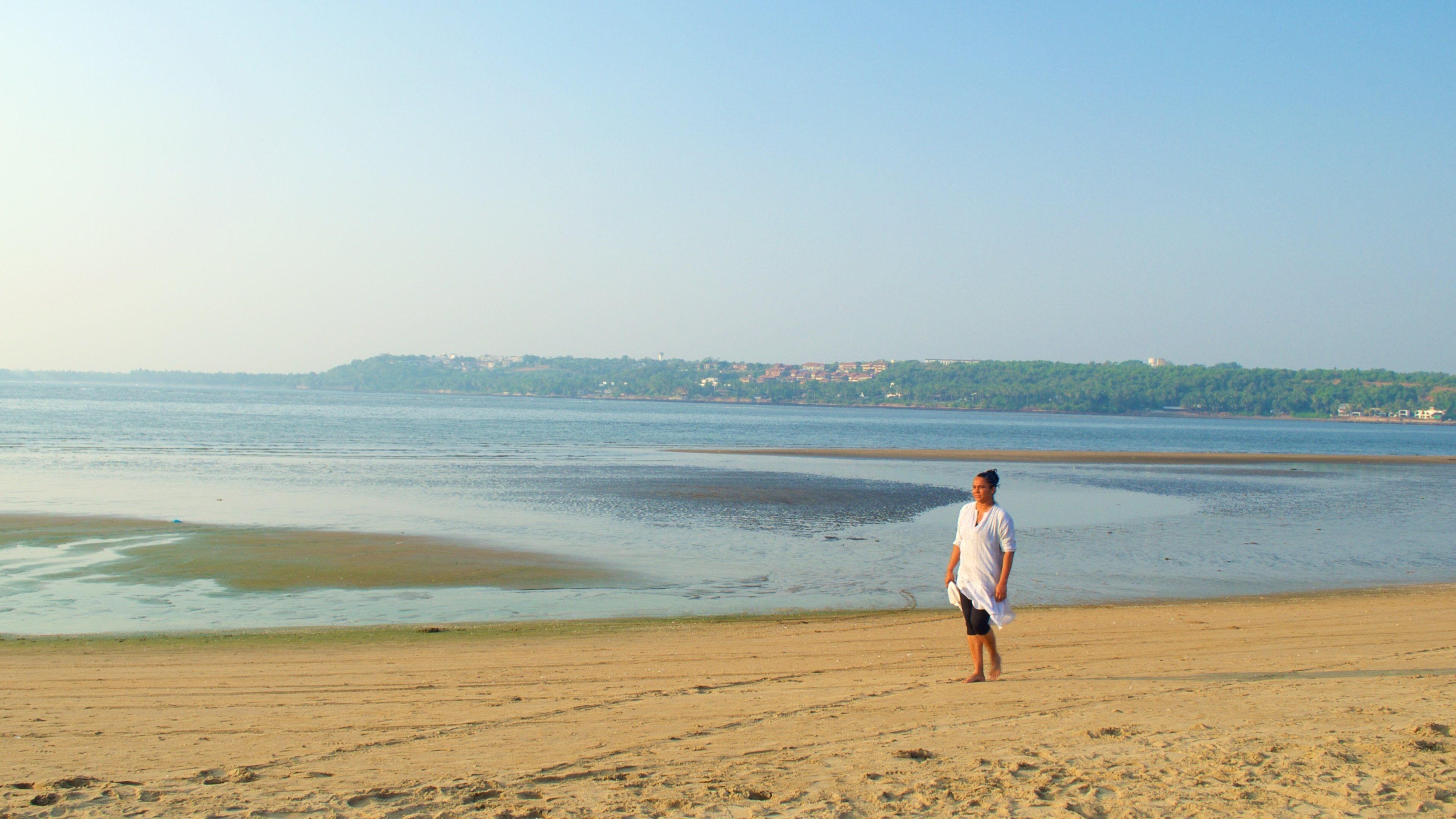 A beach with a person standing on the sand, a calm sea, and a distant shoreline with buildings.
