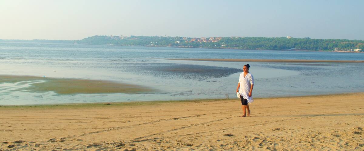 A beach with a person standing on the sand, a calm sea, and a distant shoreline with buildings.