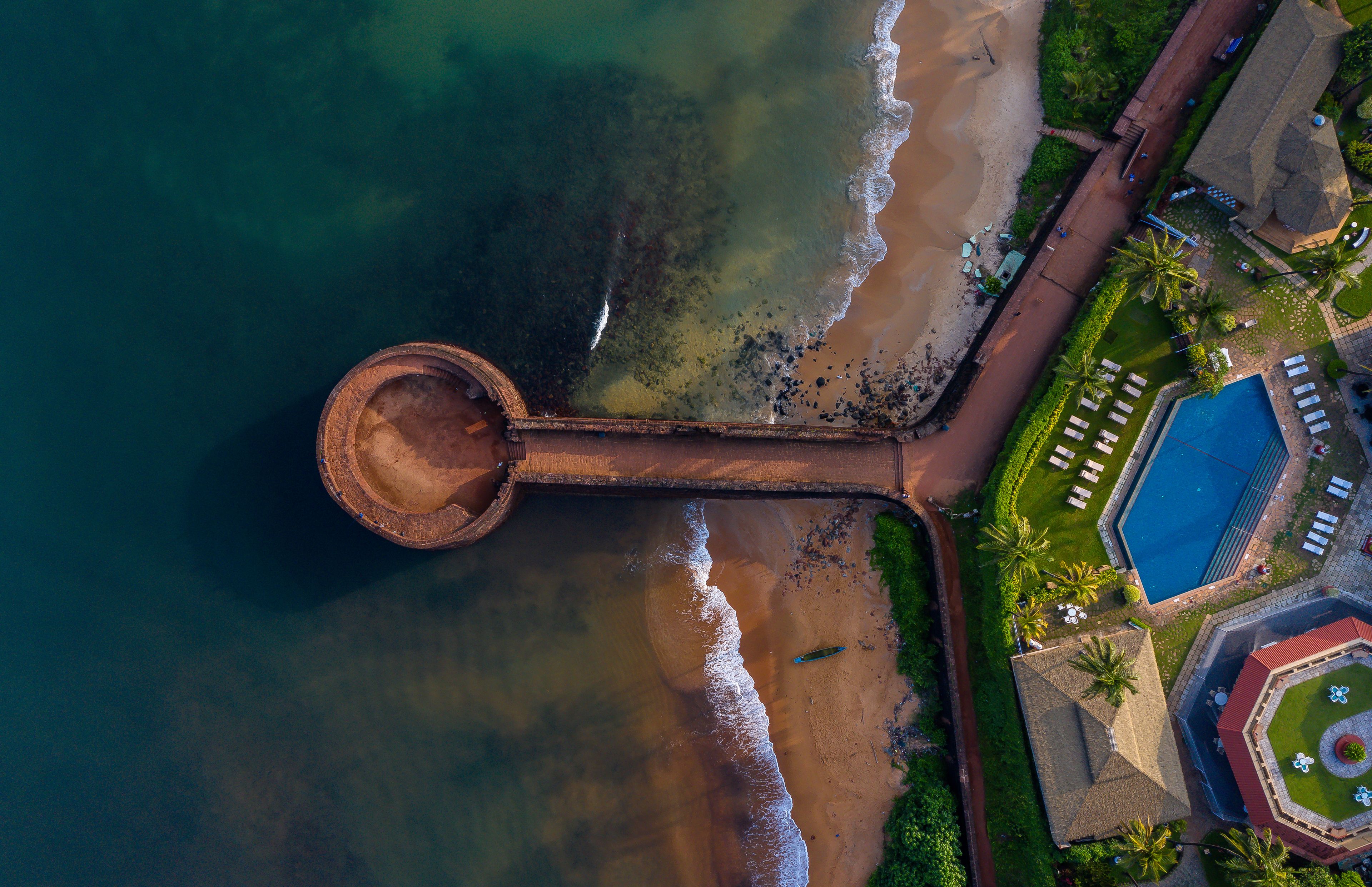 Aerial view of the historic Fort Aguada stone bastion extending into the turquoise sea with a sandy beach and a luxury resort pool Candolim, Goa, India.