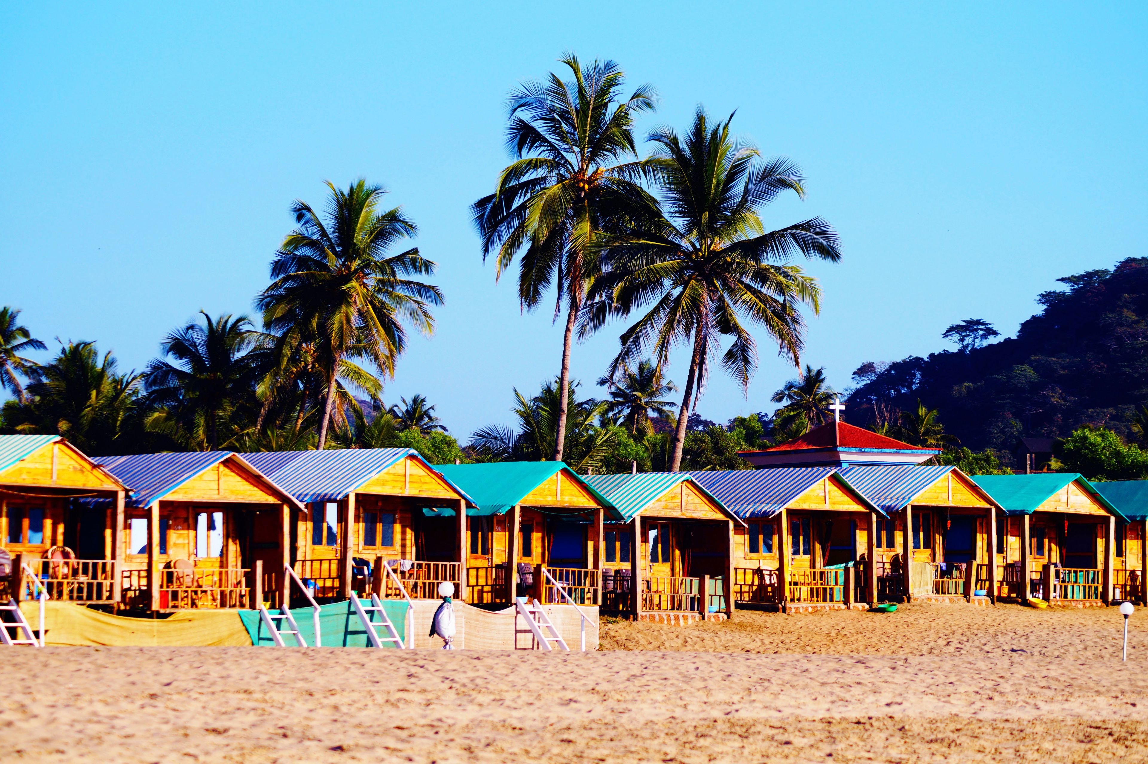 A row of wooden beach huts with blue roofs, palm trees, and a sandy beach.