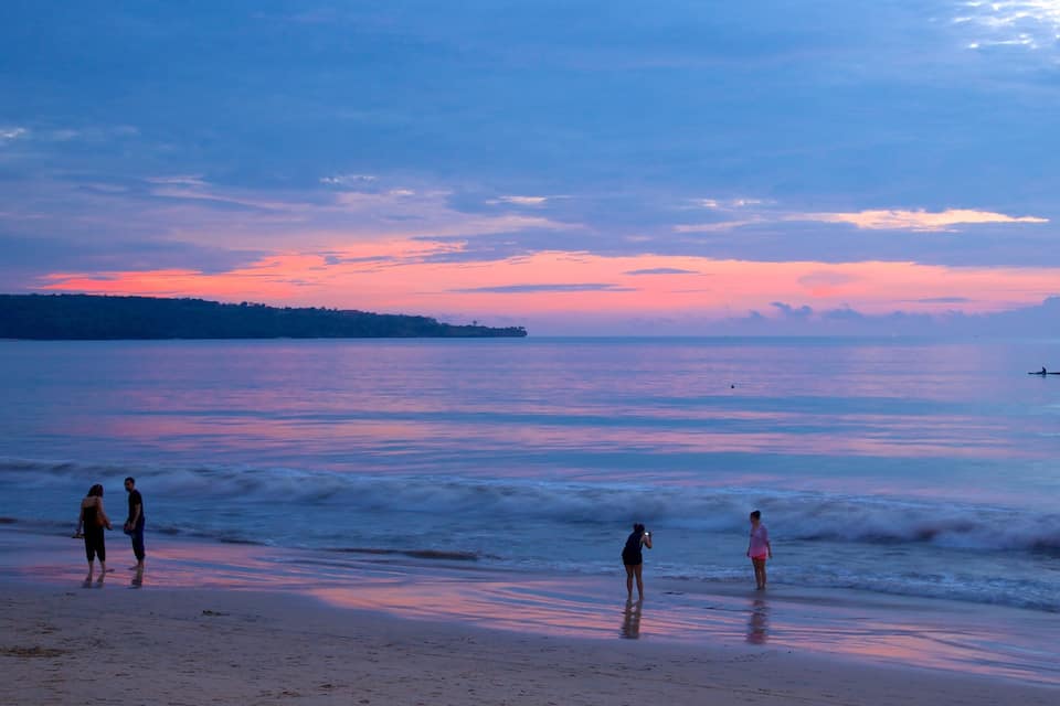 A beach at sunset with calm waters, a distant shoreline, and a few people walking along the shore.