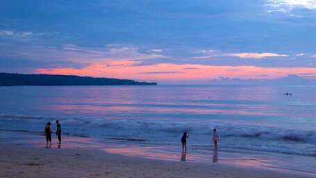 Ein Strand bei Sonnenuntergang mit ruhigem Wasser, einer entfernten Küstenlinie und einigen Menschen, die am Ufer entlanggehen.