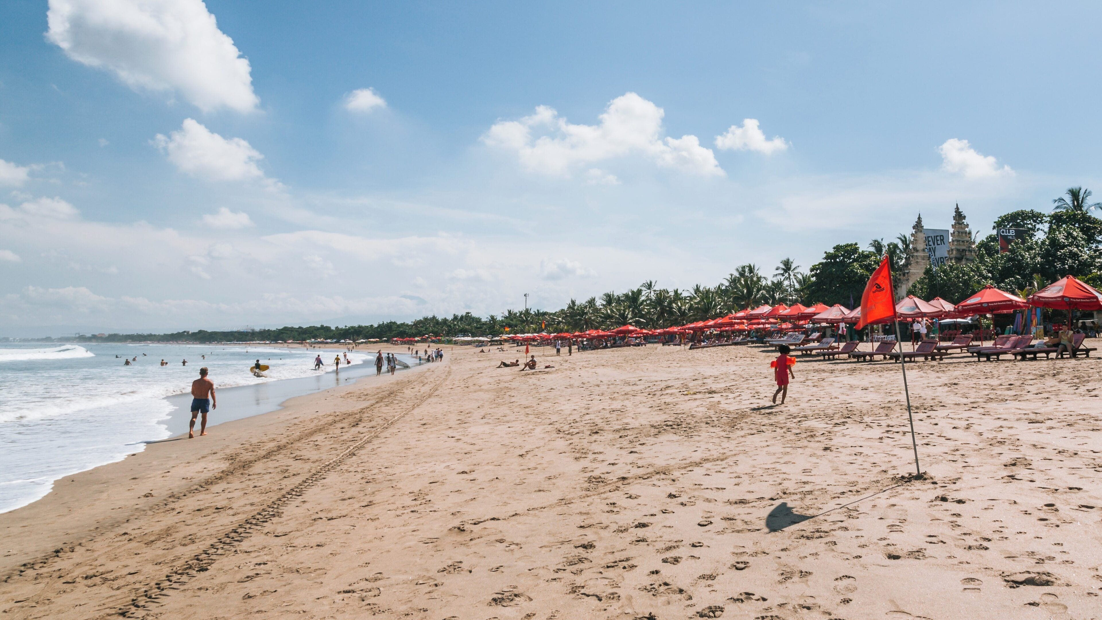 Visitors enjoy sunny day at Kuta Beach in downtown Kuta, Bali, Indonesia with vibrant red umbrellas lining the shore and gentle waves