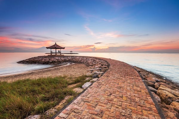 A coastal walkway with a gazebo and a small structure, leading to a calm sea under a sunset sky.