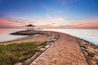 A coastal walkway with a gazebo and a small structure, leading to a calm sea under a sunset sky.