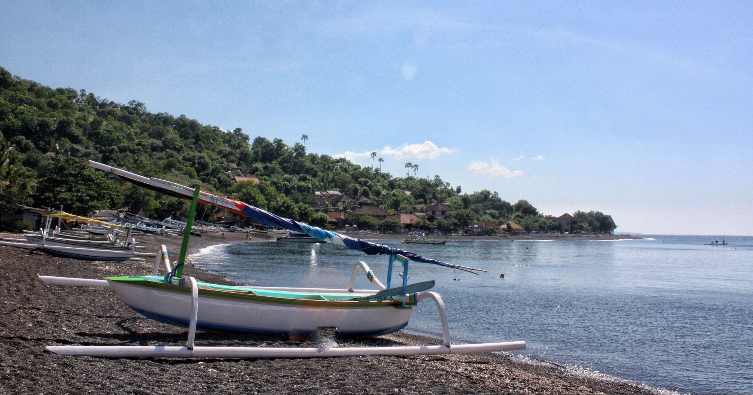 A beach with a row of boats on the shore, a clear sky, and a forested hillside in the background.