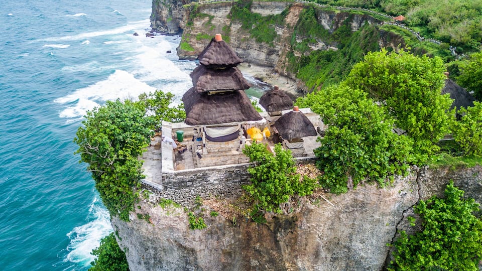 A cliffside temple complex with thatched roofs overlooking the ocean.