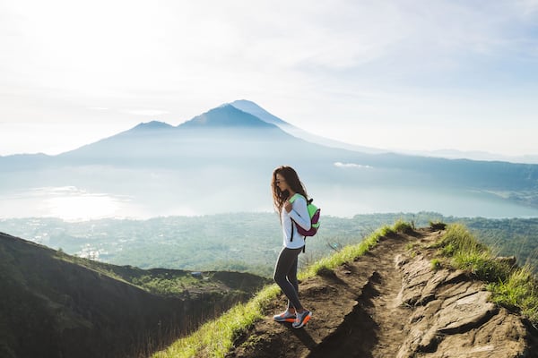 A hiker on a mountain trail with a view of a volcano and a lake.