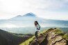 A hiker on a mountain trail with a view of a volcano and a lake.