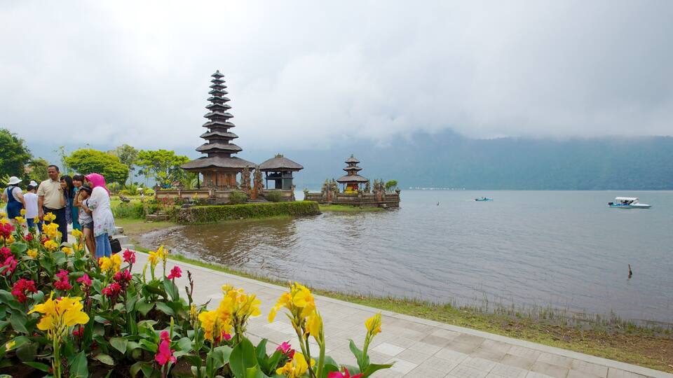 A traditional Balinese temple with tiered roofs by a calm lake, surrounded by vibrant flowers and a group of people.