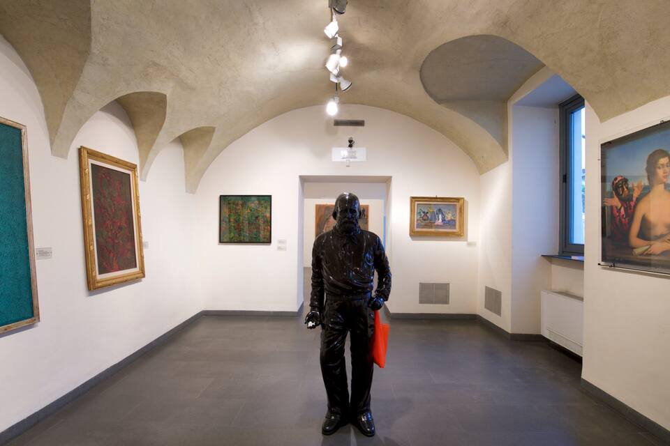 Une galerie d’art où l’on peut admirer une sculpture représentant un homme vêtu de noir, tenant un objet orange, et où les murs sont ornés de tableaux encadrés.