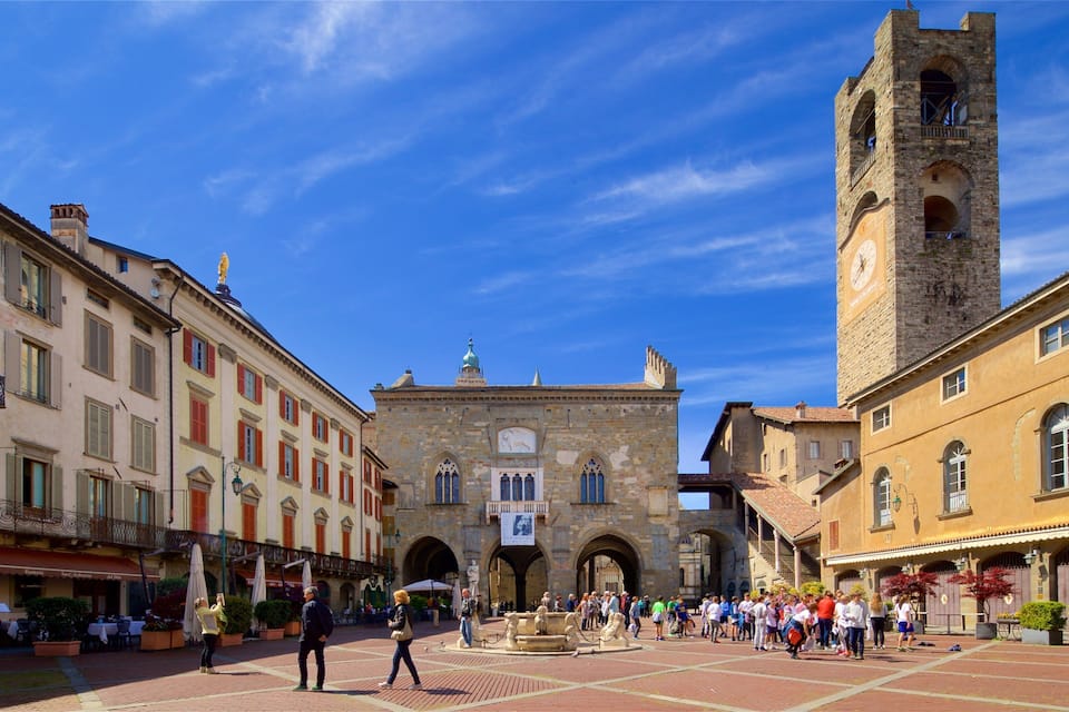 A historic square with a prominent tower, a fountain, and surrounding buildings with balconies and outdoor seating.