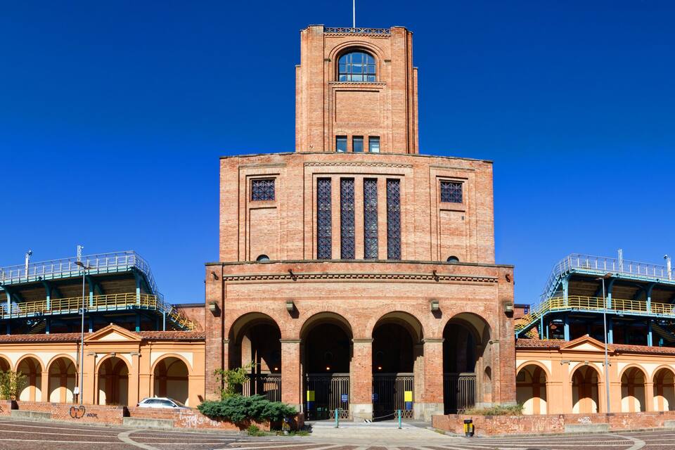 Un grand bâtiment en briques, doté d’une tour centrale et d’entrées en arc de cercle, entouré d’un stade avec des rampes bleues.