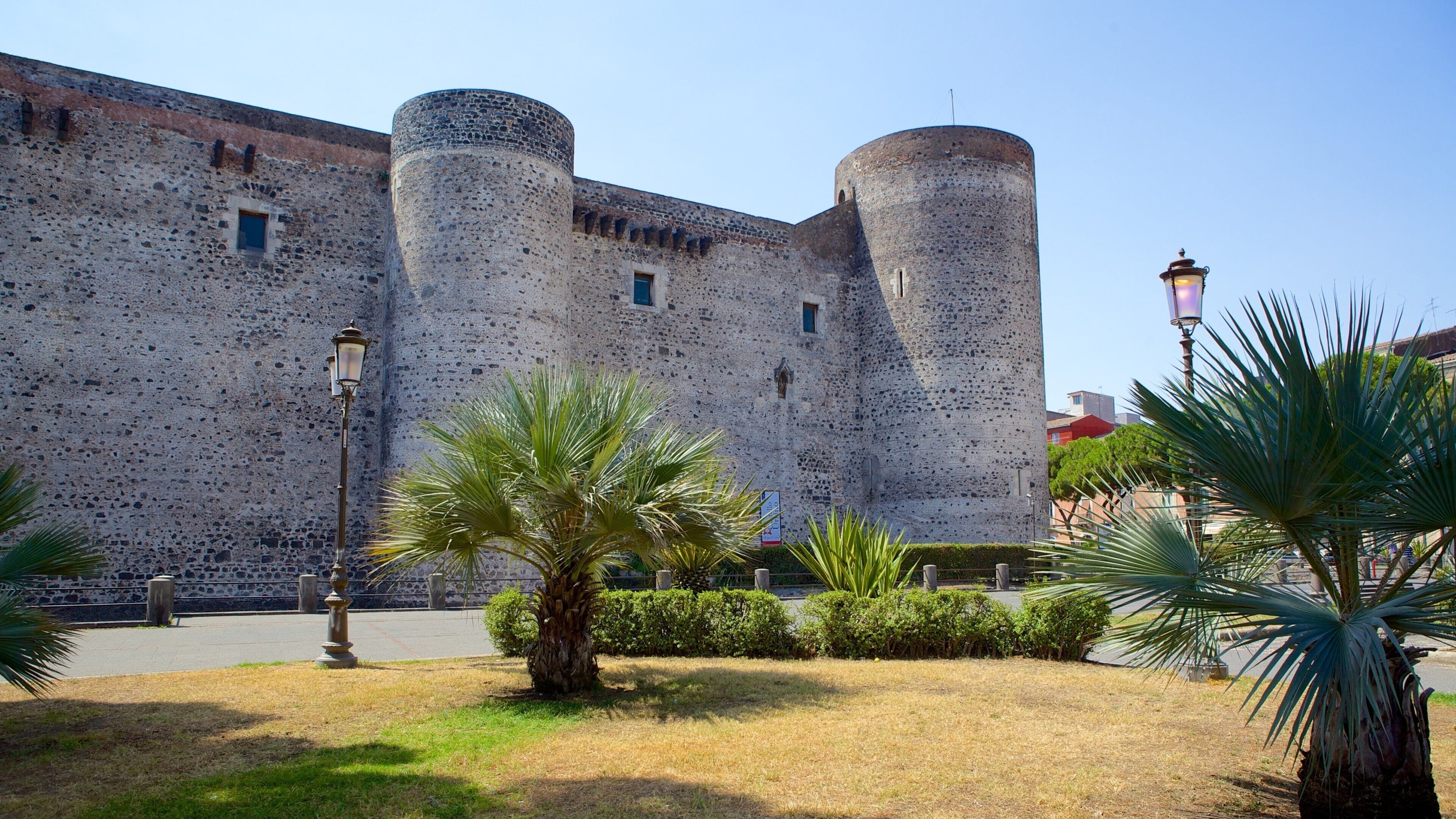 A historic stone castle with two round towers and a well-maintained garden with palm trees.