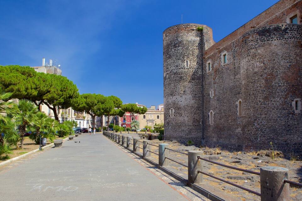 Un mur de pierre d’époque, surmonté d’une tour cylindrique, un sentier pavé bordé d’arbres et, au loin, des bâtiments traditionnels.