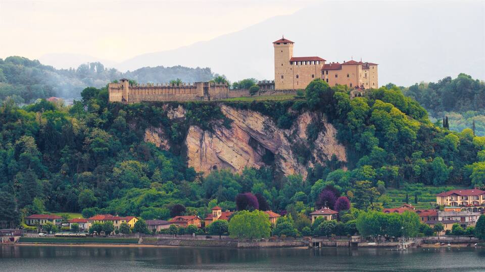 A castle on a cliff overlooking a lake, surrounded by lush greenery and residential buildings.