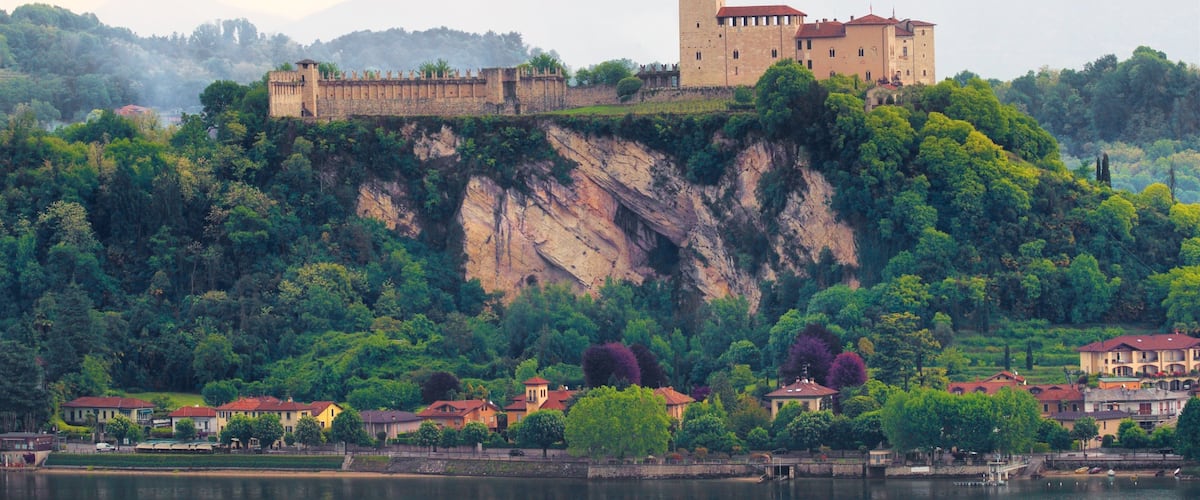A castle on a cliff overlooking a lake, surrounded by lush greenery and residential buildings.