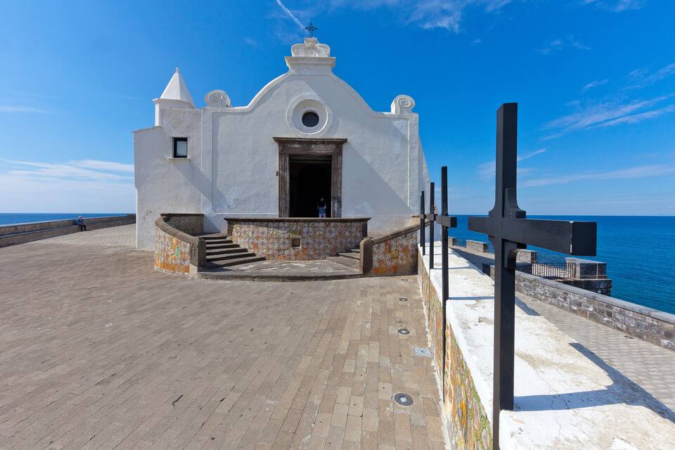 A white church with a bell tower and a cross on a paved walkway by the sea.