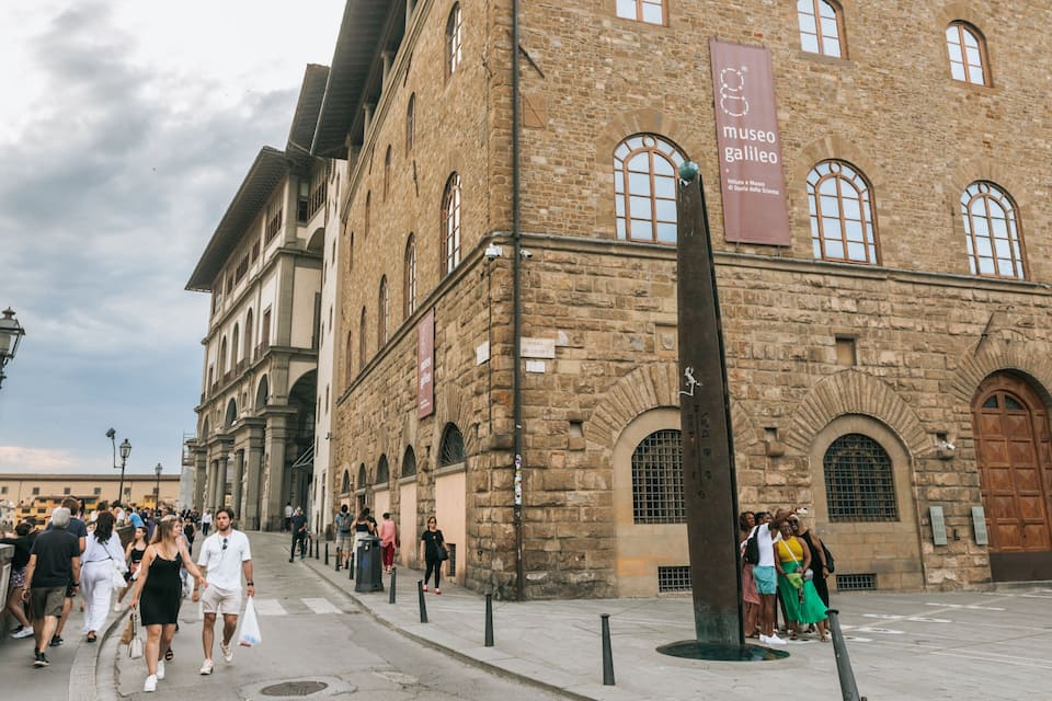 A historic building with stone walls and arched windows, a crowd of people, and a street lamp.