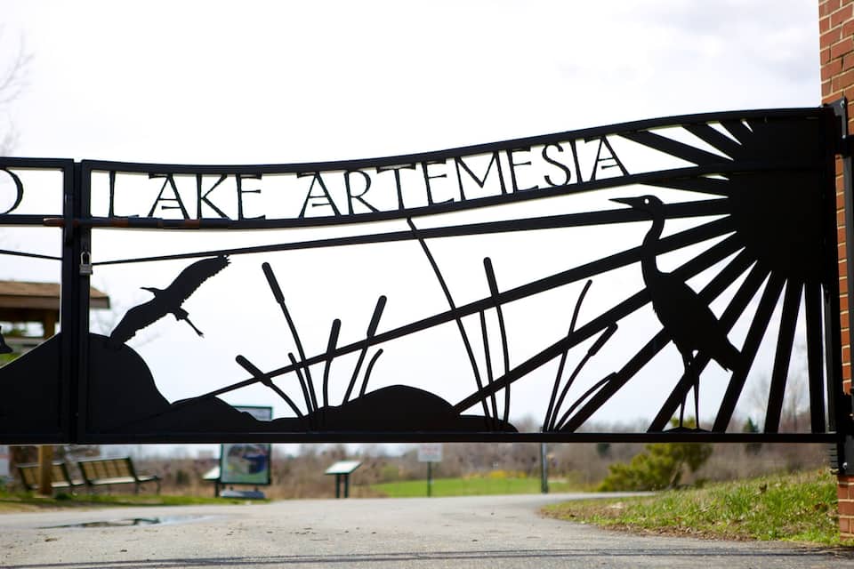 A metal gate with the words "TO LAKE ARTEMISIA" and silhouettes of a bird and a fox.