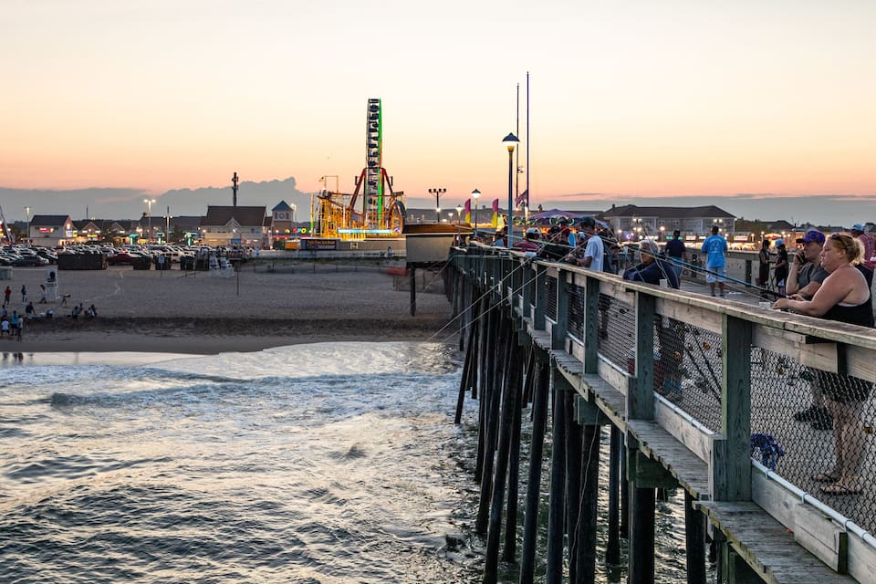 A pier with people on it, a beach, and a roller coaster in the background.