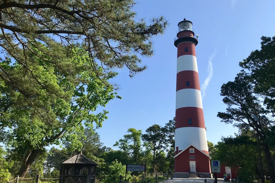 A red and white lighthouse with a small adjacent building, surrounded by trees and a clear sky.