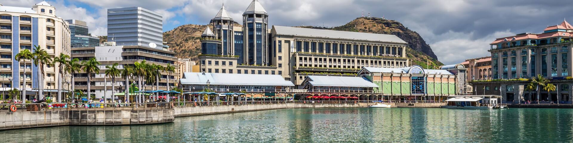 A waterfront cityscape with a prominent building featuring a tower, palm trees, and a hill in the background.