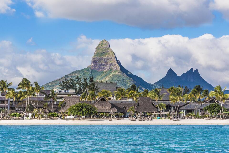 A beach with thatched-roof buildings and palm trees, a mountain in the background, and clear blue water.