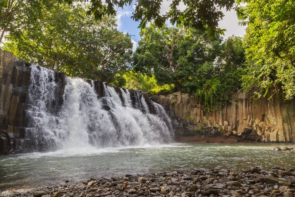 A waterfall cascading over a rocky cliff into a river, surrounded by dense green foliage.