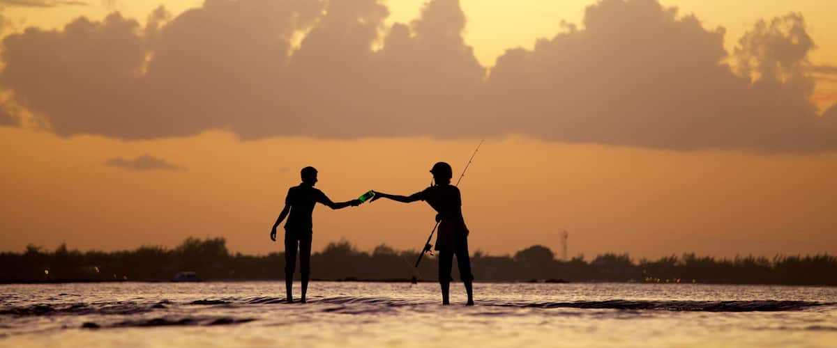 Two people fishing at sunset with a calm sea and a cloudy sky.