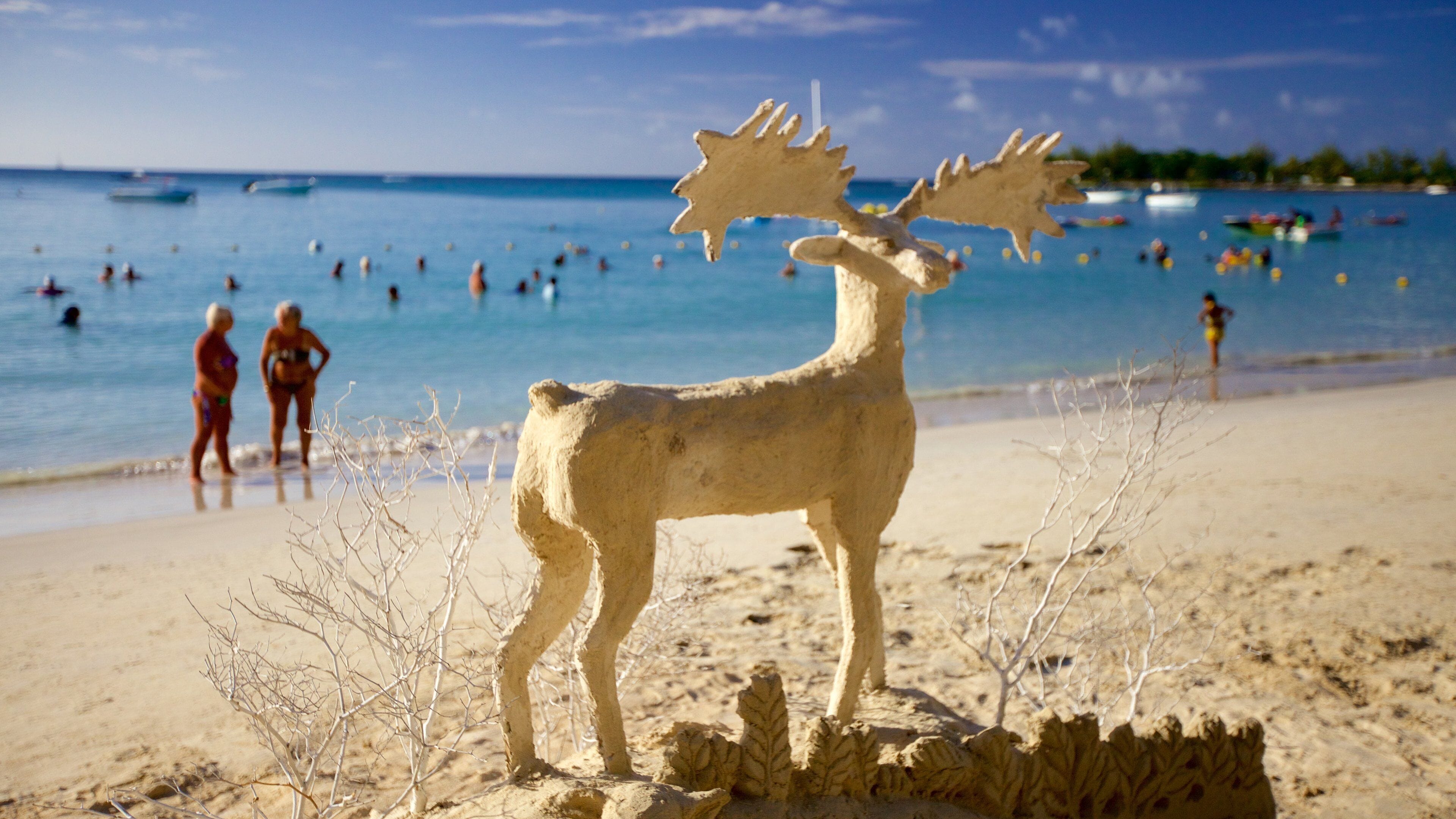 A sand sculpture of a moose on a beach with people in the water and boats in the background.