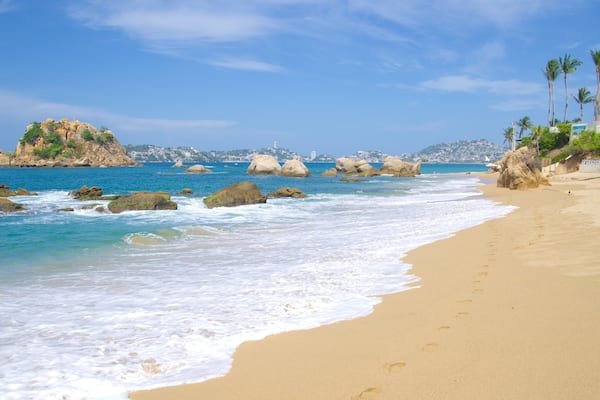 Ein sandiger Strand mit klarem, blauem Wasser, großen Felsen und Palmen.