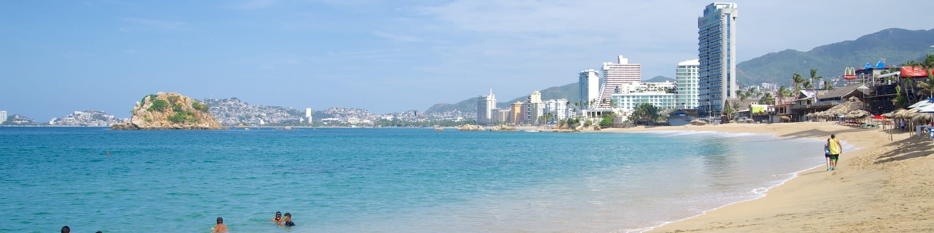 Una playa con aguas azules cristalinas, una orilla arenosa y un paisaje urbano al fondo.