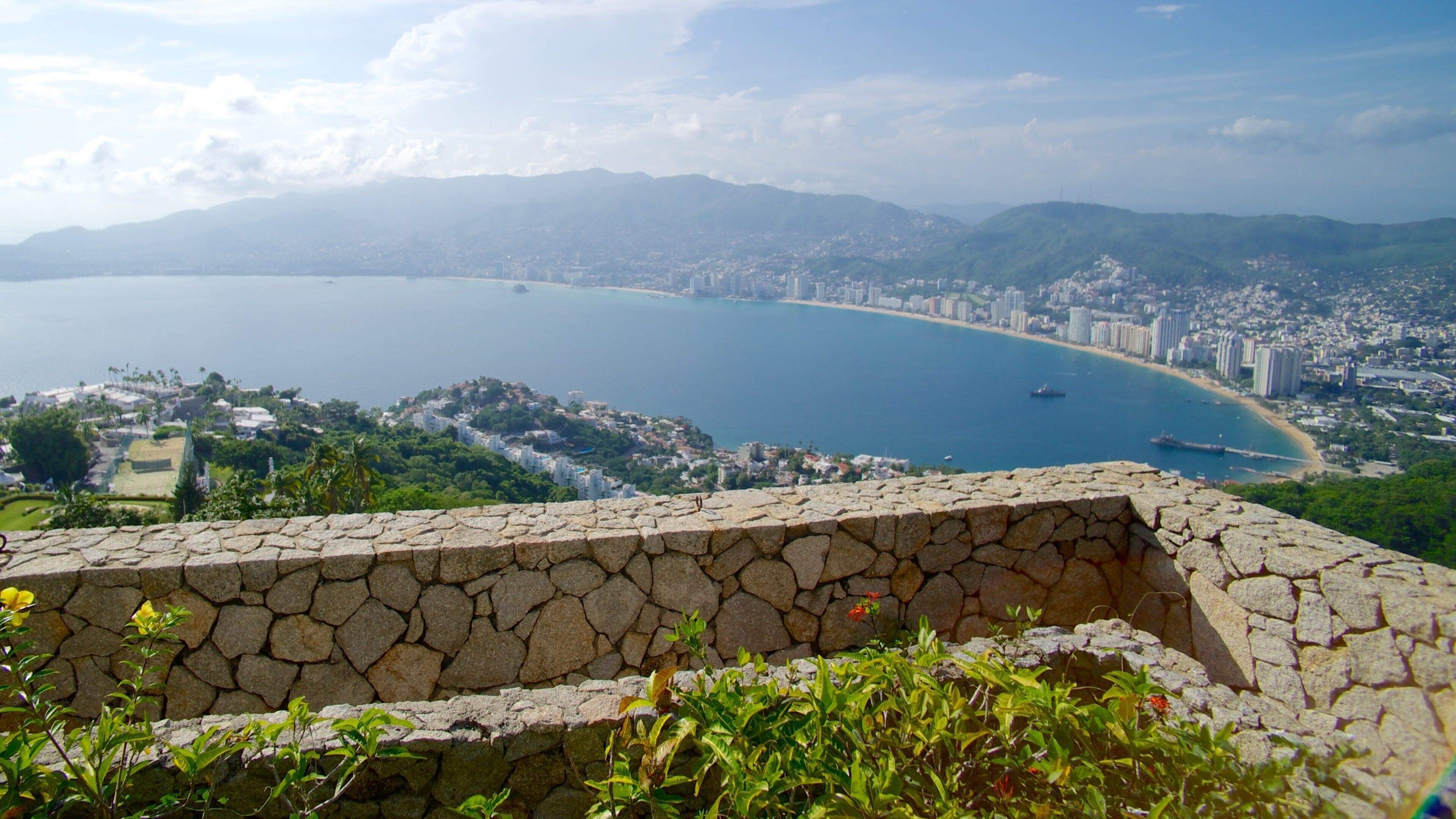 A coastal city with a large bay, a stone wall in the foreground, and mountains in the background.