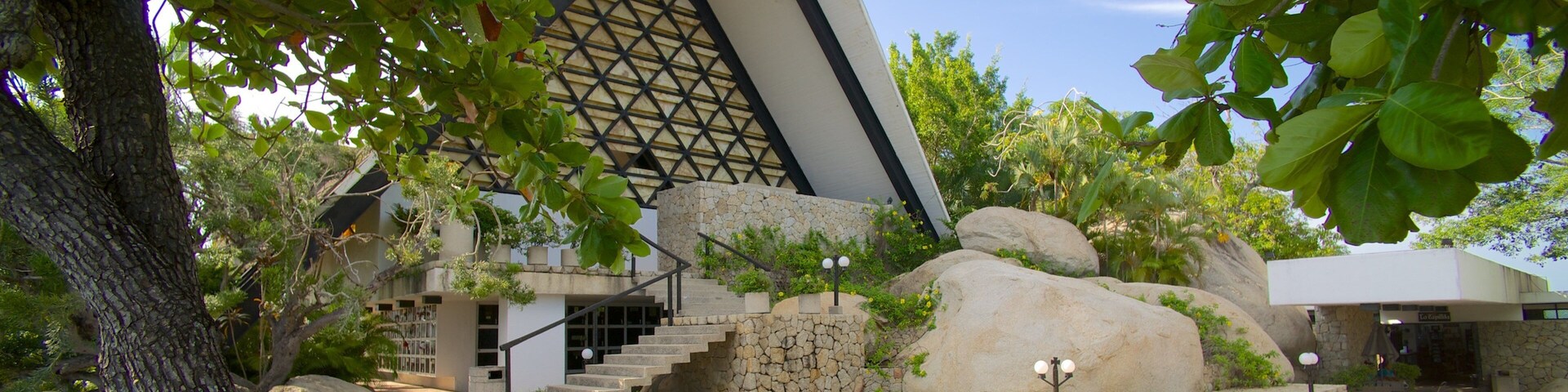 Um edifício moderno com fachada geométrica, cercado por pedras grandes e vegetação exuberante.