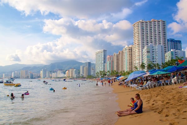 A crowded beach with people, boats, and tall buildings in the background.