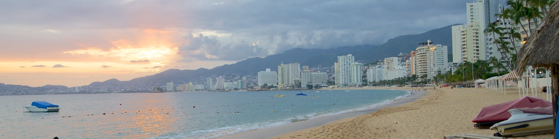 Uma praia com um horizonte urbano ao fundo, um barco na água e pessoas caminhando pela orla.