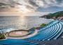 A seaside amphitheater with blue steps and a circular stage, overlooking the ocean.