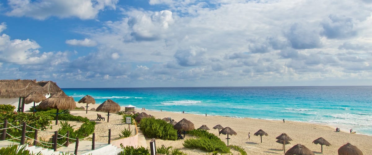 Une plage avec des parasols en chaume, une eau turquoise et limpide, et un ciel partiellement nuageux.