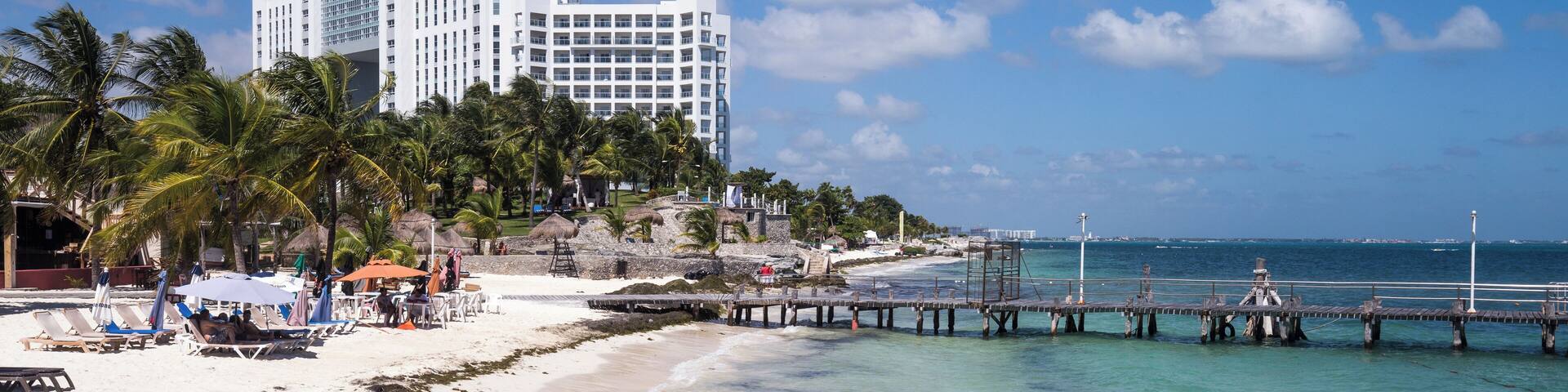 A beach with a hotel, palm trees, and a pier extending into the ocean.