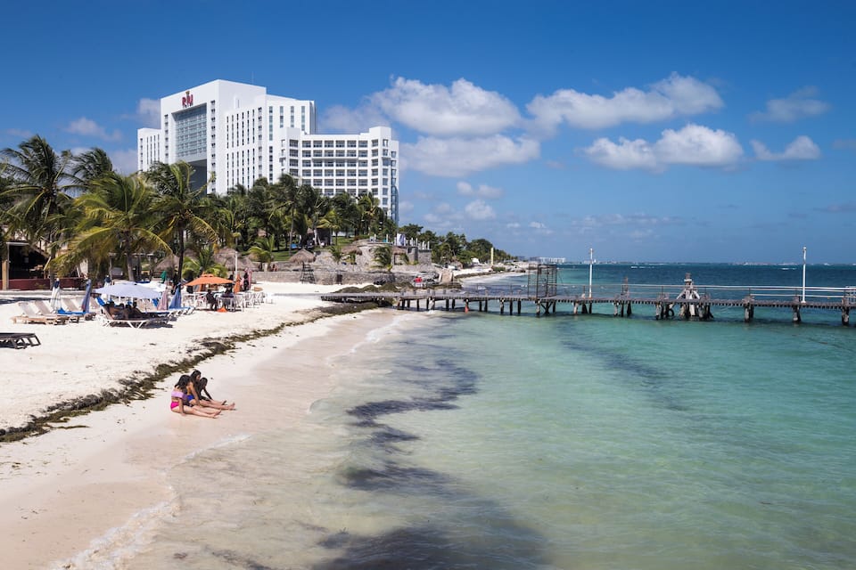A beach with a hotel, palm trees, and a pier extending into the ocean.