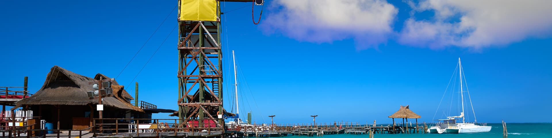 Un muelle con un edificio de techo de paja, una estructura metálica alta y un velero en aguas turquesas.