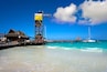 A pier with a thatched-roof building, a tall metal structure, and a sailboat in turquoise waters.