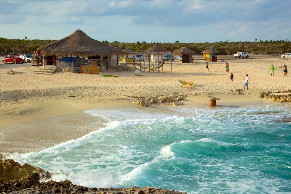 A beach with thatched-roof huts, a rocky shoreline, and people walking along the sand.
