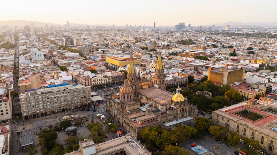 A cityscape with a prominent cathedral featuring two tall spires and a golden dome, surrounded by dense urban buildings and a clear sky.
