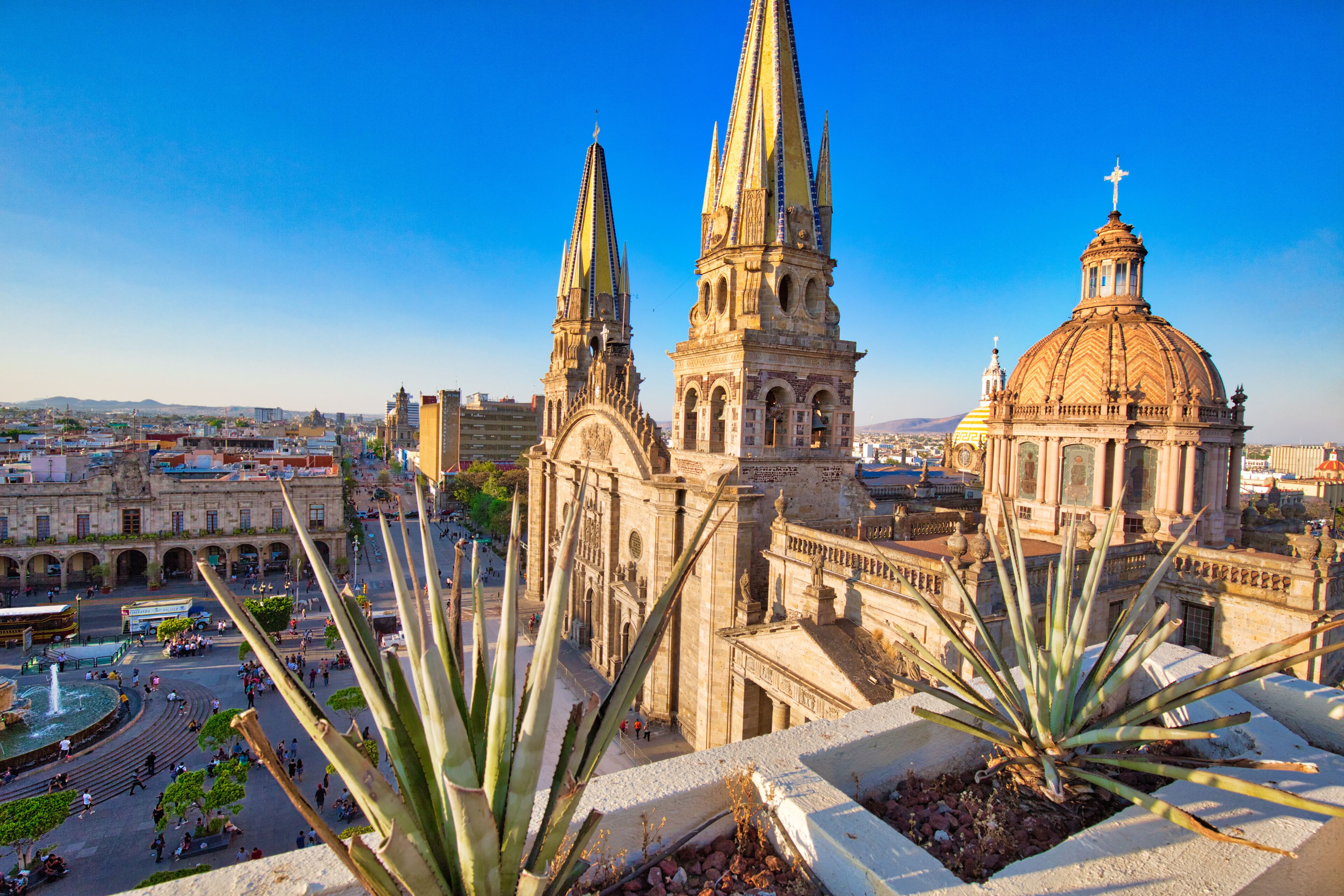 A cathedral with twin spires and a large dome, surrounded by a plaza with people and a fountain.