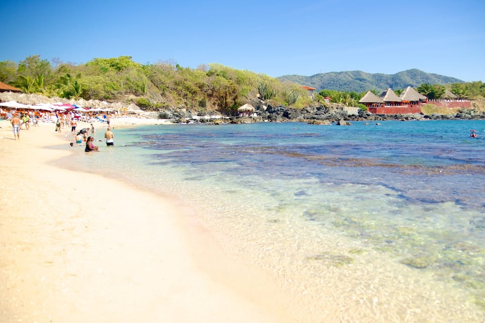 A beach with clear shallow waters, a sandy shore, and thatched-roof huts in the background.