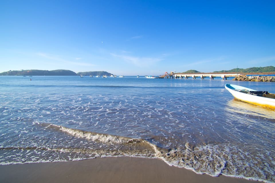 A calm beach with gentle waves, a pier extending into the water, and boats docked nearby.